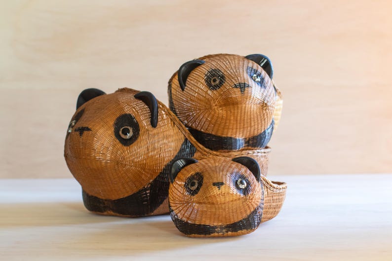 May include: Three woven panda baskets, one stacked on top of the other. The top basket is brown with black eyes and a black nose. The middle basket is brown with black eyes and a black nose. The bottom basket is brown with black eyes and a black nose. The baskets are made of woven material and have a natural, rustic look.