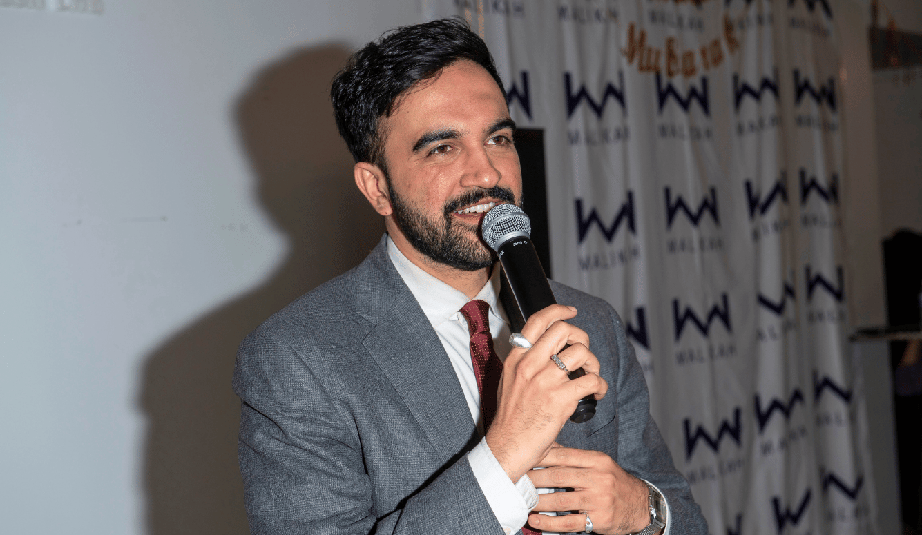 Mamdani is a young light-skinned Indian-looking man in a suit. He speaks into a microphone. Mamdani is a young light-skinned Indian-looking man in a suit. He speaks into a microphone.