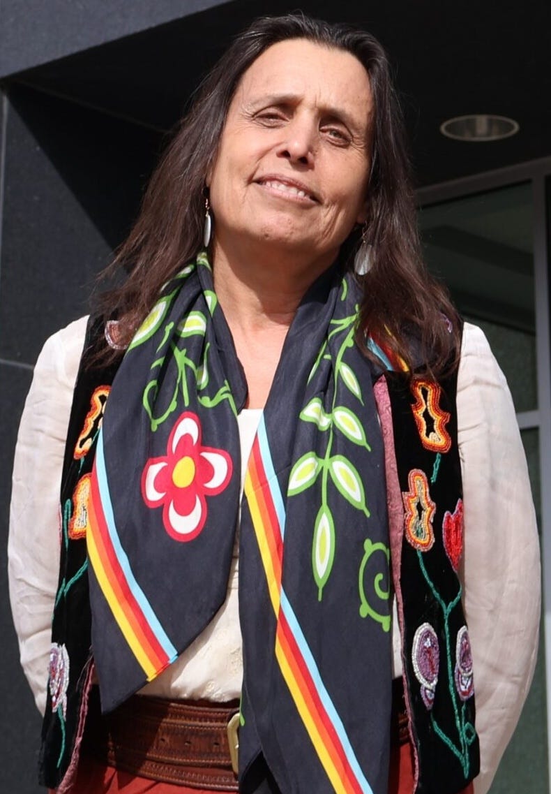Photo of Winona, looking at the camera with long brown hair, her head a little tilted to the side, wearing a black scarf with a multicolored flower pattern on it, with tan olive brown skin, a slight smile, and an off white shirt below the scarf.