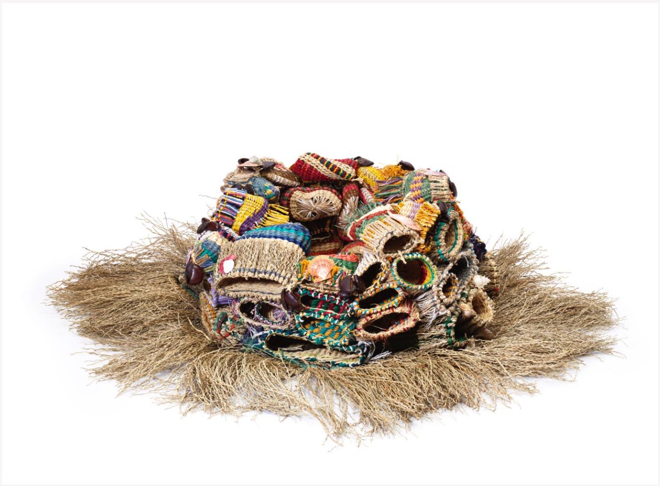 A large, sculptural woven artwork composed of layered, coiled, and tubular forms made from natural fibres and brightly coloured materials. The piece features a dense cluster of interwoven shapes in reds, yellows, greens, and blues, with repeating circular openings and textured surfaces. It sits on a base of loose, straw-like fibres that spread outward, evoking the organic form of a reef or marine structure.