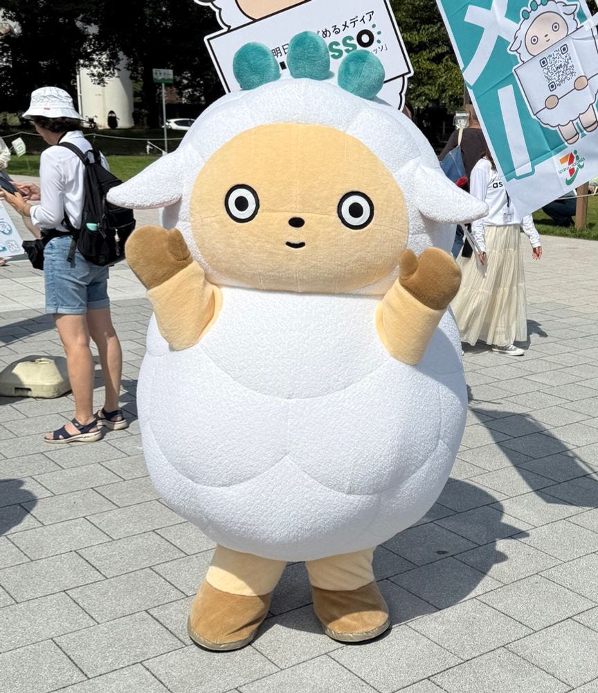 A sheep mascot with three green circles on its head and a vacant stare poses on a pavement with its hands raised.
