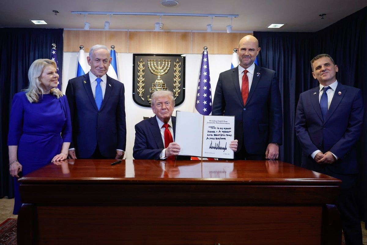 President Donald J. Trump seated at a wooden desk in a formal room with wooden panels and flags signs a guest book while holding a pen. Standing beside him are Prime Minister Benjamin Netanyahu in a dark suit and blue tie, US Ambassador David Friedman in a gray suit and red tie, and First Lady Melania Trump in a blue dress. The setting includes Israeli and American flags on the wall, ornate lighting fixtures, and a professional diplomatic atmosphere at the Knesset.
