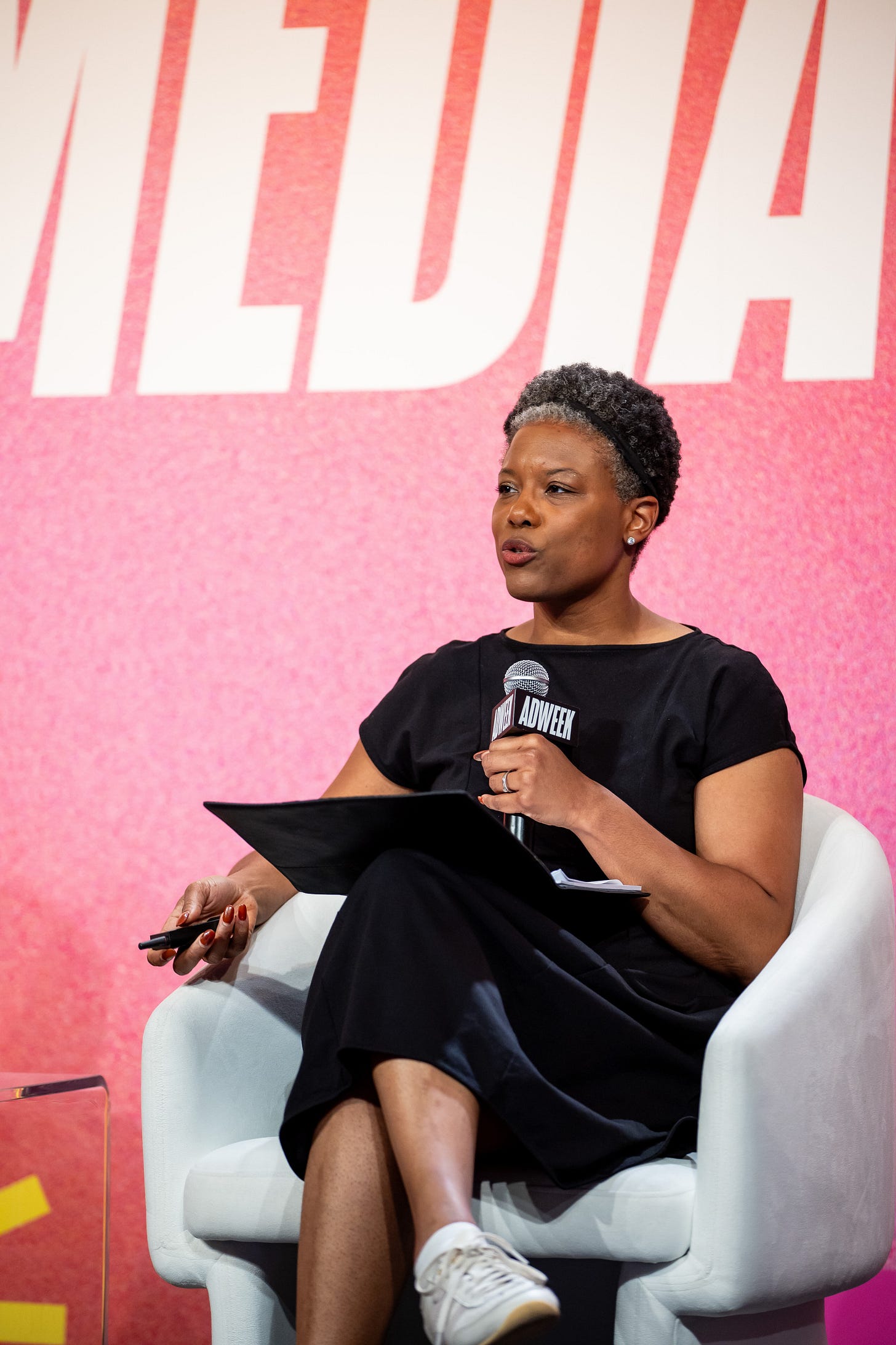 An image of Brittany Hennessy, a woman with brown skin and short black hair. She is speaking into a microphone against a hot pink background.