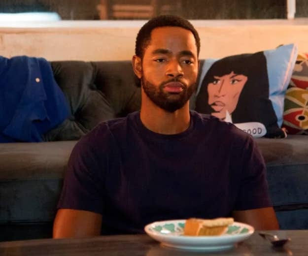 A man sitting on a couch looking pensive, wearing a dark t-shirt, with a plate of food on the coffee table in front of him. A still from the TV show Insecure.