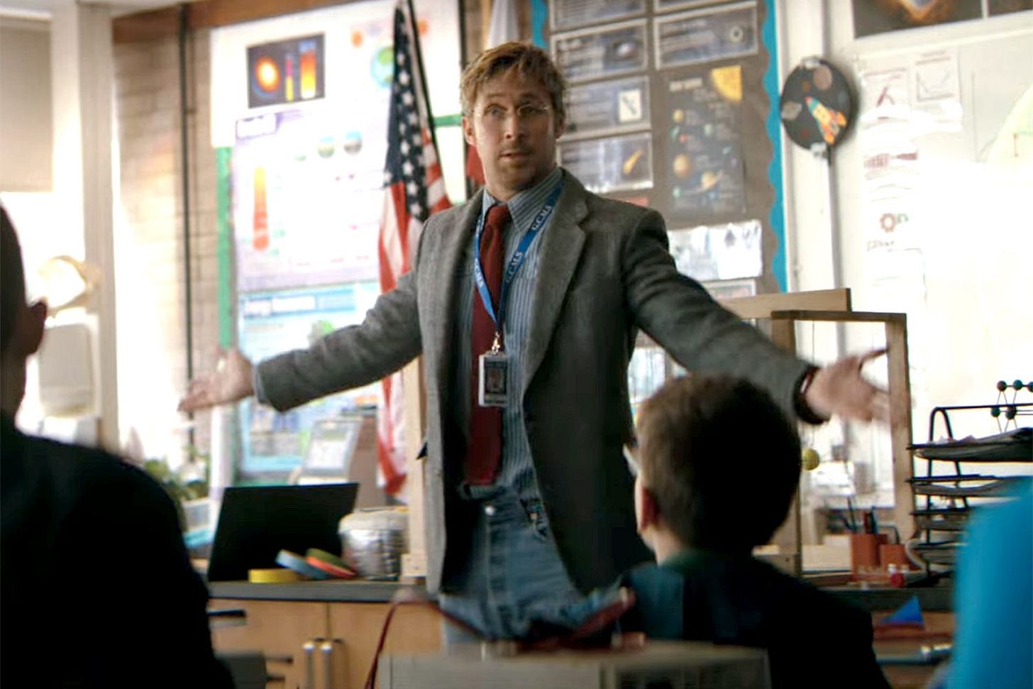 A science teacher standing at the front of a classroom with arms outstretched, speaking to students, with science posters and an American flag in the background. A science teacher standing at the front of a classroom with arms outstretched, speaking to students, with science posters and an American flag in the background.