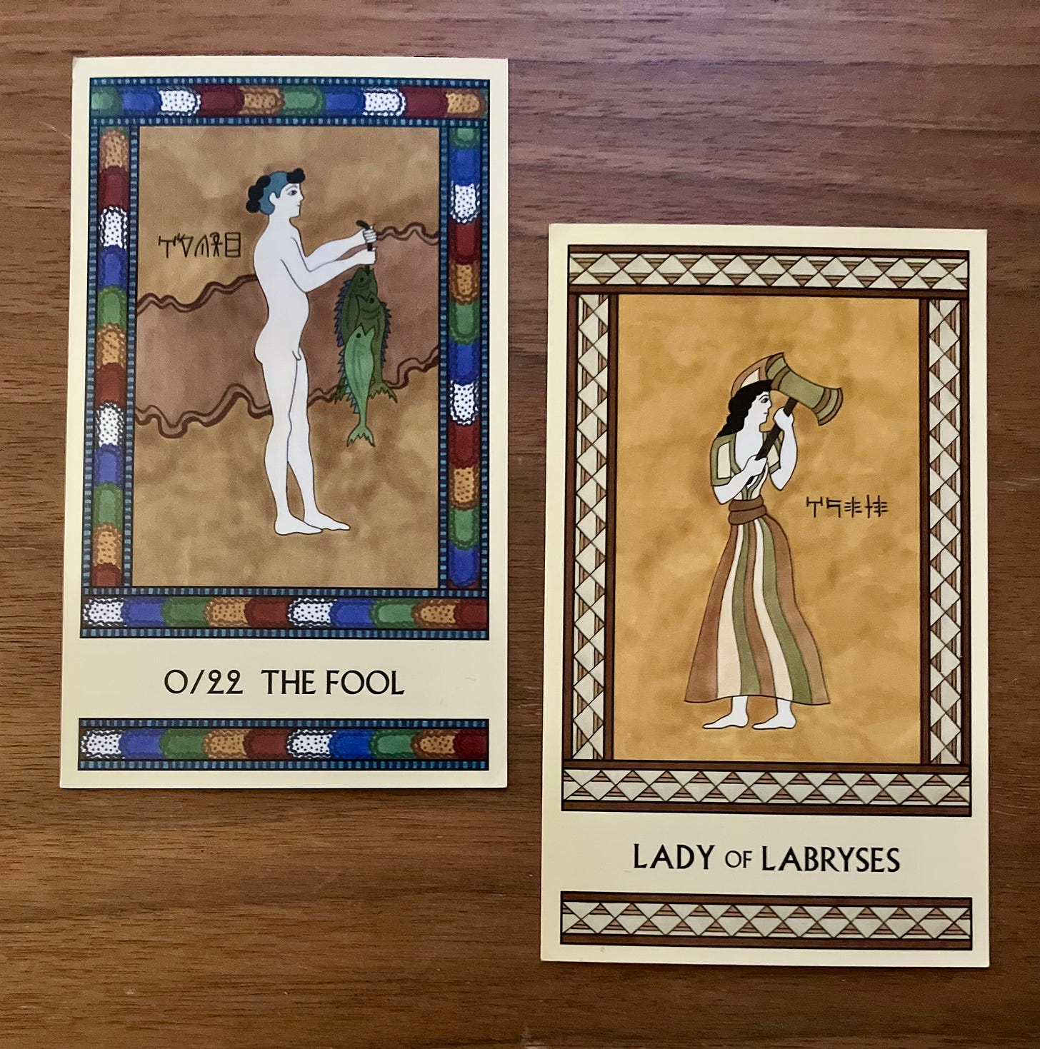 Two Minoan Tarot cards on a wood background. The Fool has a multicolor border and shows a naked boy facing right, holding a string of just-caught fish. The Lady of Labryses is in shades of gold and cream. It shows a woman in Minoan dress, facing left but looking right over her shoulder and carrying a labrys.