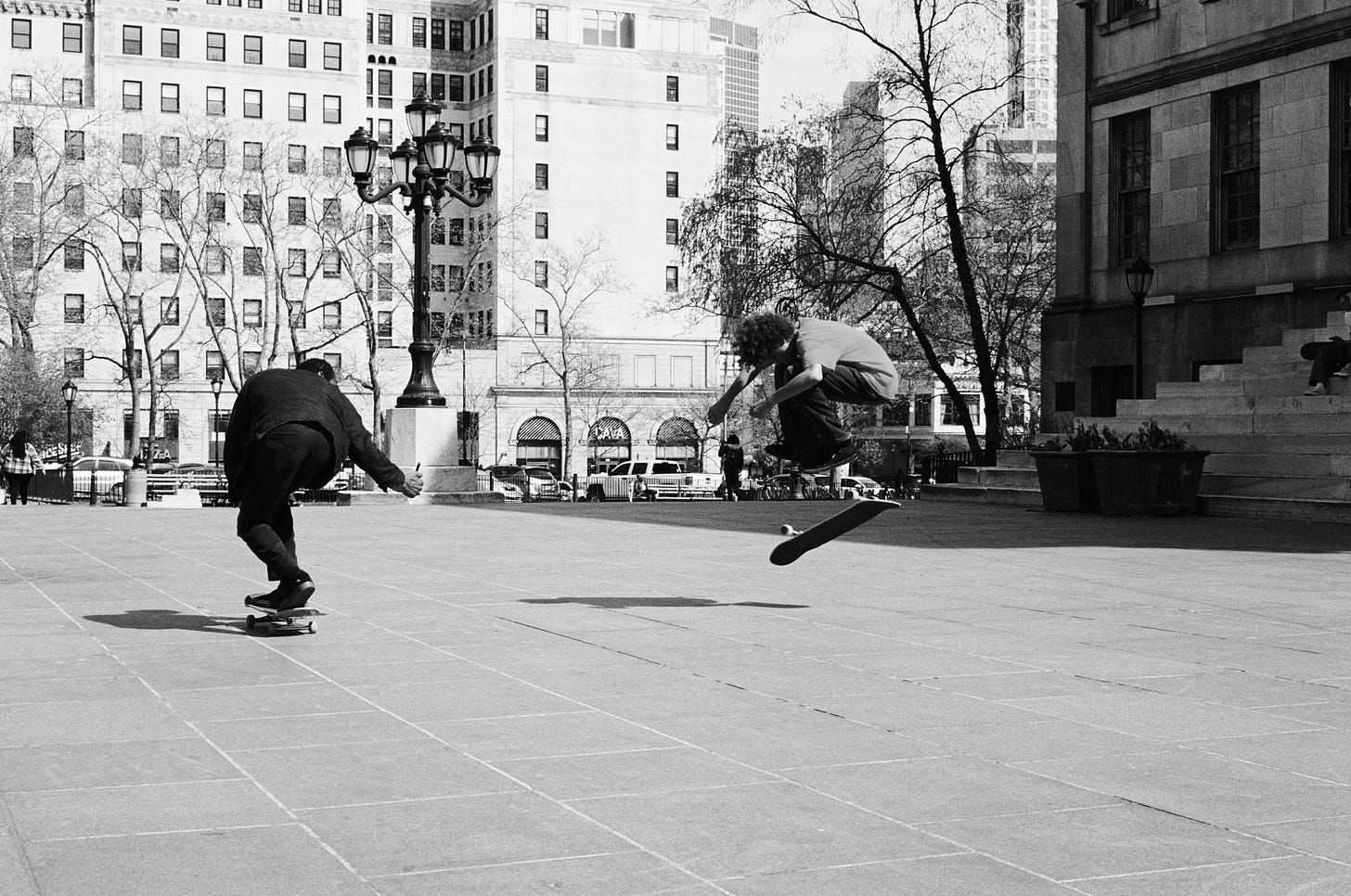 A father skateboards while filming his son mid-air doing a trick on his skateboard in a public plaza
