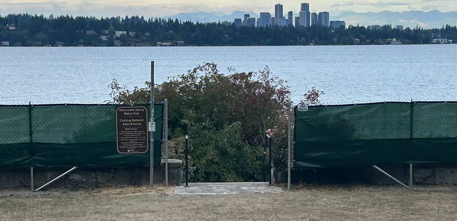 A view facing Lake Washington from Denny Blaine Park in Seattle, showing new chain-link fencing with green tarps dividing the park’s nude and clothed areas. A brown sign reads “Clothing Optional: East of Fence,” with city park rules listed below. The skyline of Bellevue is visible across the lake in the distance.
