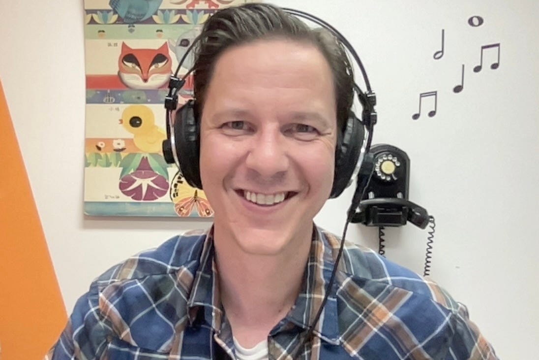 Eric Powell wearing headphones in his studio. Hes wearing a plad shirt. In the background is an illustrated picture of animals and flowers.