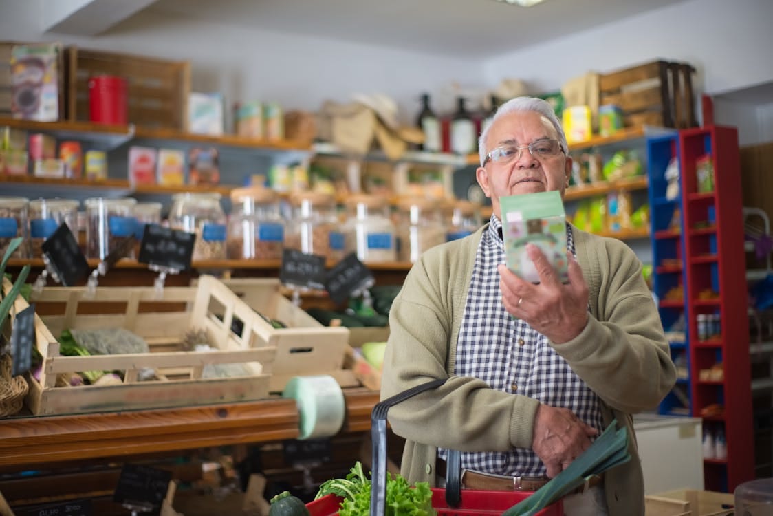 Free Elderly man examines product in grocery store aisle, representing daily shopping routine. Stock Photo
