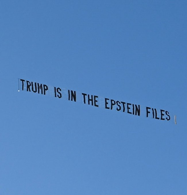 A white banner with black text reading TRUMP IS IN THE EPSTEIN FILES flies in a clear blue sky, positioned horizontally and captured from below against the horizon. A white banner with black text reading TRUMP IS IN THE EPSTEIN FILES flies in a clear blue sky, positioned horizontally and captured from below against the horizon.