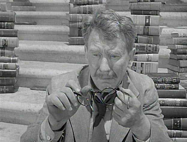 black and white image of a man sitting in front of piles of books and looking down at his broken glasses