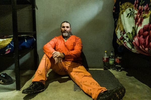 A man in orange prison garb sits on the floor of a cell beside a bunk, with a few plastic bottles and a piece of floral-print fabric next to him as well.