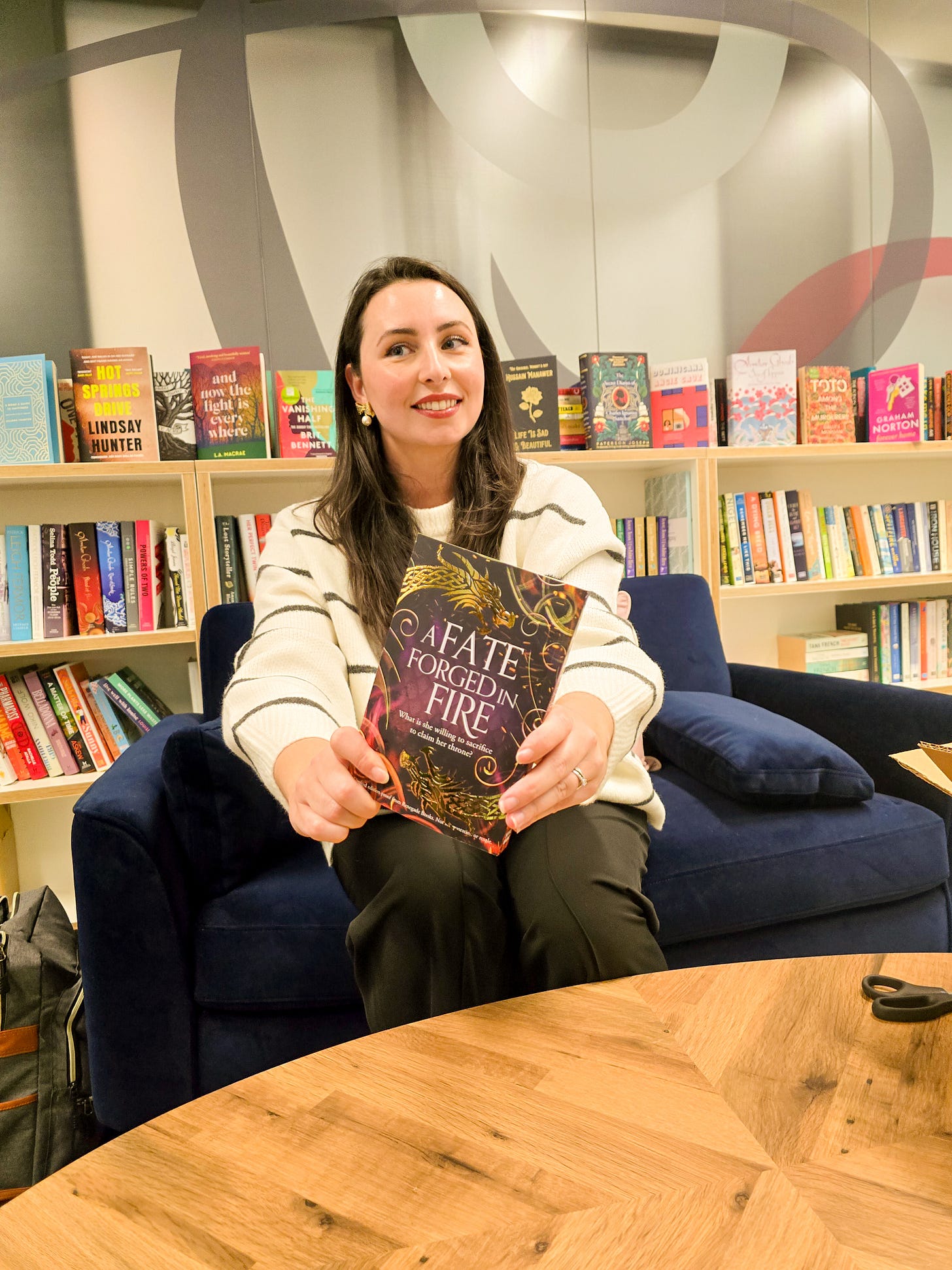Hazel sits in front of a bookshelf holding a copy of her fantasy romance novel titled ‘A FATE FORGED IN FIRE’. The cover is purple with gold foiled celtic dragons. She wears a white and black striped top and smiles while looking away from the camera