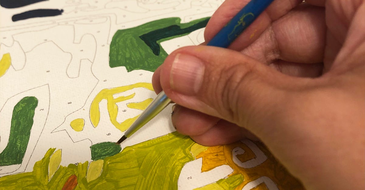 Photo of an unfinished paint by number showing a close up view of a hand with a blue paint brush painting in a green patch of colour. Other areas of the paint by number show patches of green and yellow with empty areas showing the numbers.