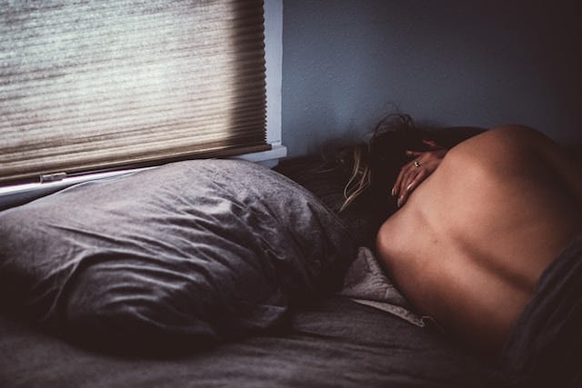A woman lying in bed, facing away from the camera, her hand resting on her neck. Her posture expresses deep sadness, as if she's grieving after a painful loss.