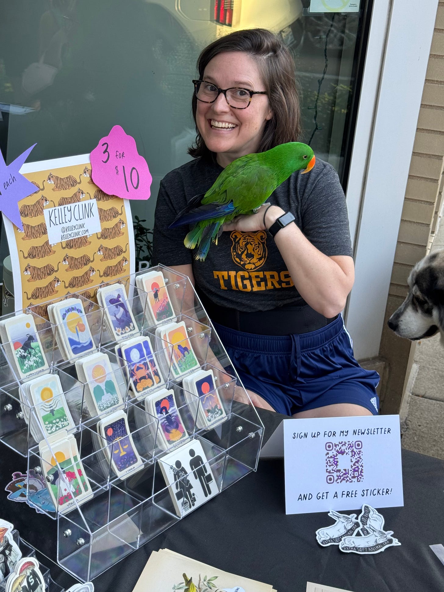 a picture of Kelley smiling wide next to their sticker display, a small, bright green parrot resting on their left hand.