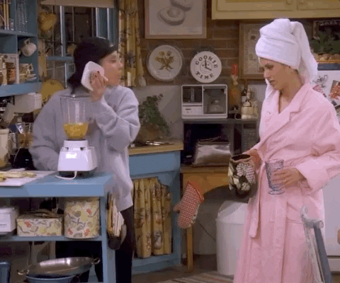 A GIF from the TV show Friends featuring Monica Geller using a blender in her kitchen while Rachel Green stands nearby in a pink bathrobe and hair towel. It illustrates the messy, real-life process of DIY skincare formulation and home beauty rituals.