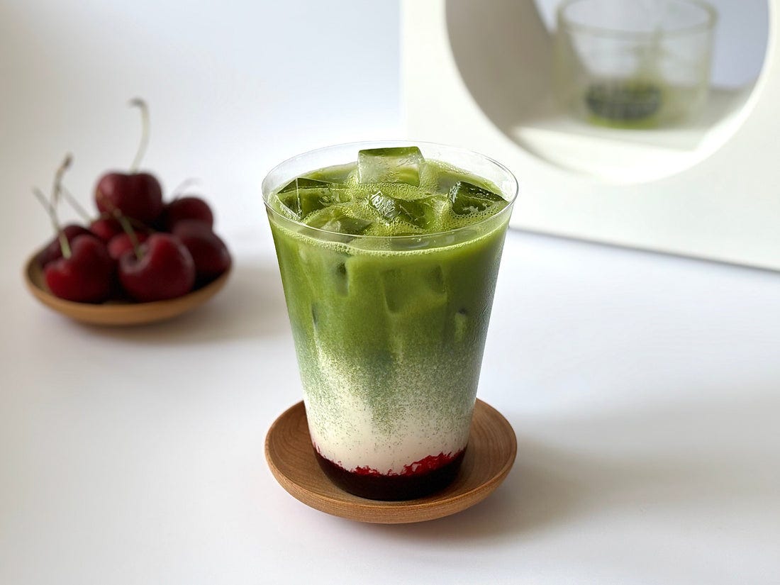 An iced cherry matcha latte with vivid green matcha, milk and deep red cherry syrup set in front of a Matcha Maker.