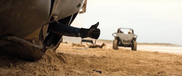 A mud-covered arm gives a thumbs-up from beneath a flipped vehicle on a sandy beach, while a stunt buggy drives away in the background.