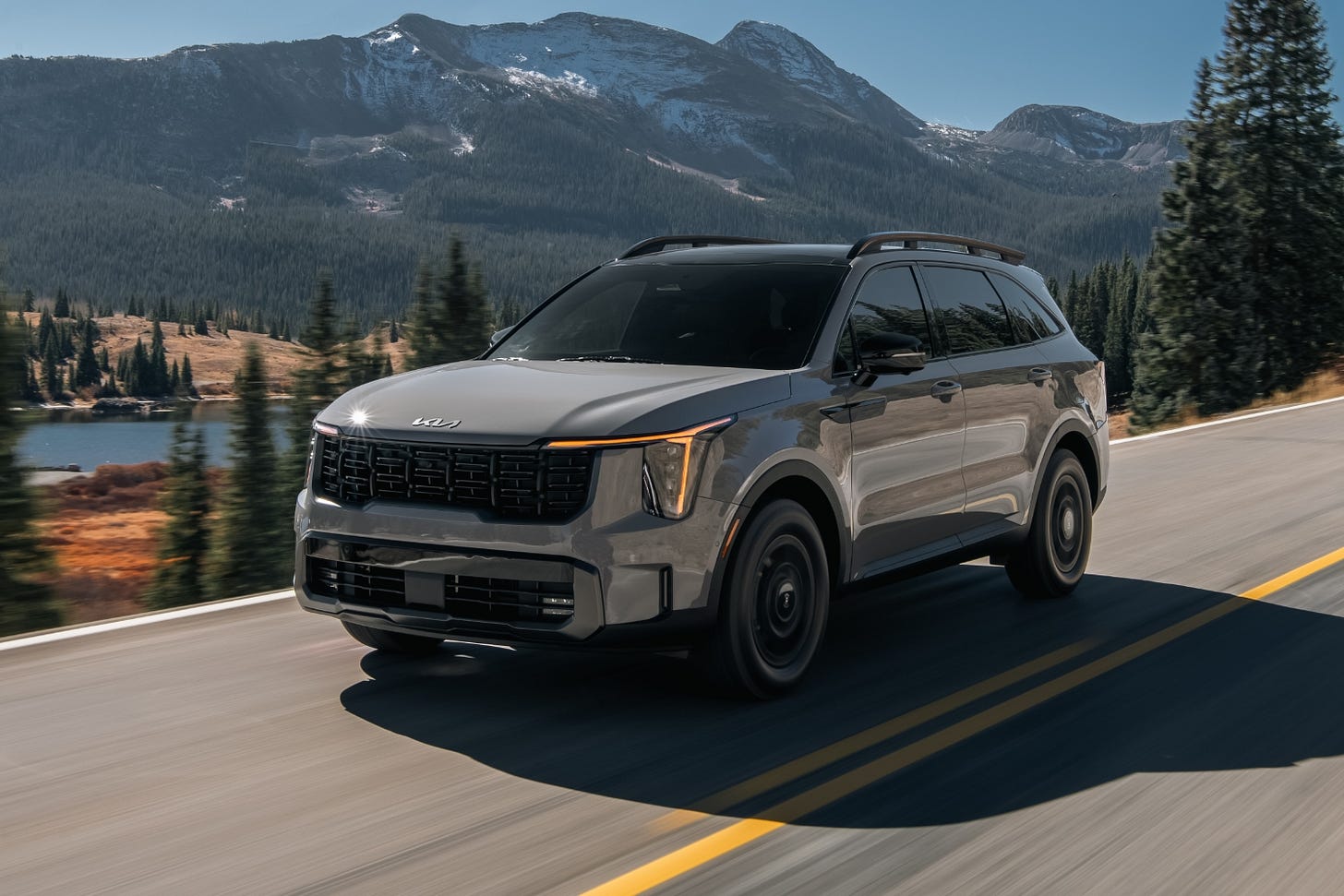 A 2024 Kia Sorento X-Pro driving on a mountain road with a lake and mountains in the background.