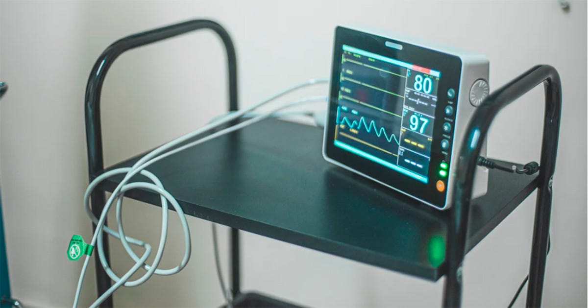 Monitor multiparamétrico hospitalar apoiado sobre um carrinho metálico preto, medindo sinais vitais, com gráficos de batimentos cardíacos e números em destaque