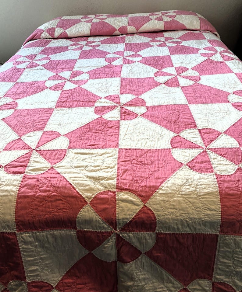 May include: A pink and white quilt with a geometric pattern. The quilt features alternating squares and circular designs in a patchwork style. The bedspread is made of a shiny fabric and is neatly arranged on a bed. May include: A pink and white quilt with a geometric pattern. The quilt features alternating squares and circular designs in a patchwork style. The bedspread is made of a shiny fabric and is neatly arranged on a bed.