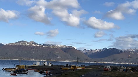 A road bordered by buildings, facing the water; a man's hands pointing through the windshield with a machine gun visible through the driver's window; a fishing dock with mountains and a town on the other side of a stretch of water.