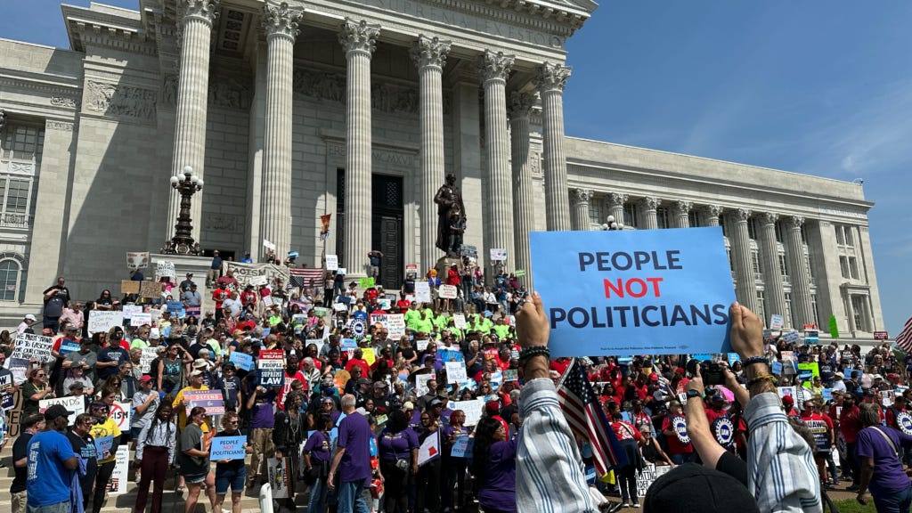 Thousands of people stand in front of the Missouri capitol building holding signs demanding an end to democracy. In the foreground hands hold a sign saying People Not Politicians. Thousands of people stand in front of the Missouri capitol building holding signs demanding an end to democracy. In the foreground hands hold a sign saying People Not Politicians.