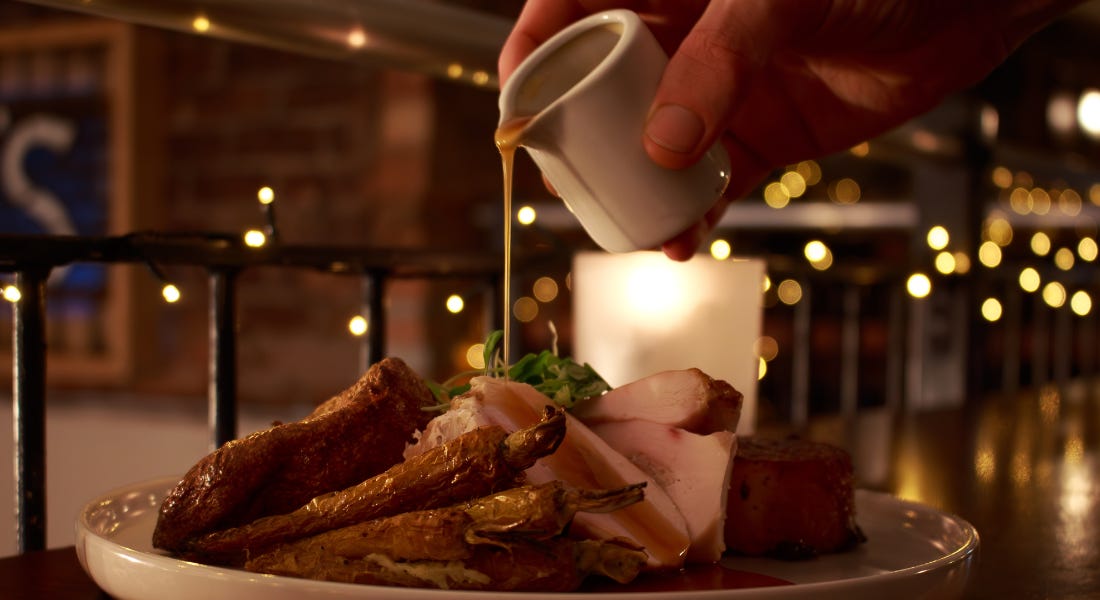 A plate of turkey roulade and vegetables with gravy being poured on it, with fairy lights in the background