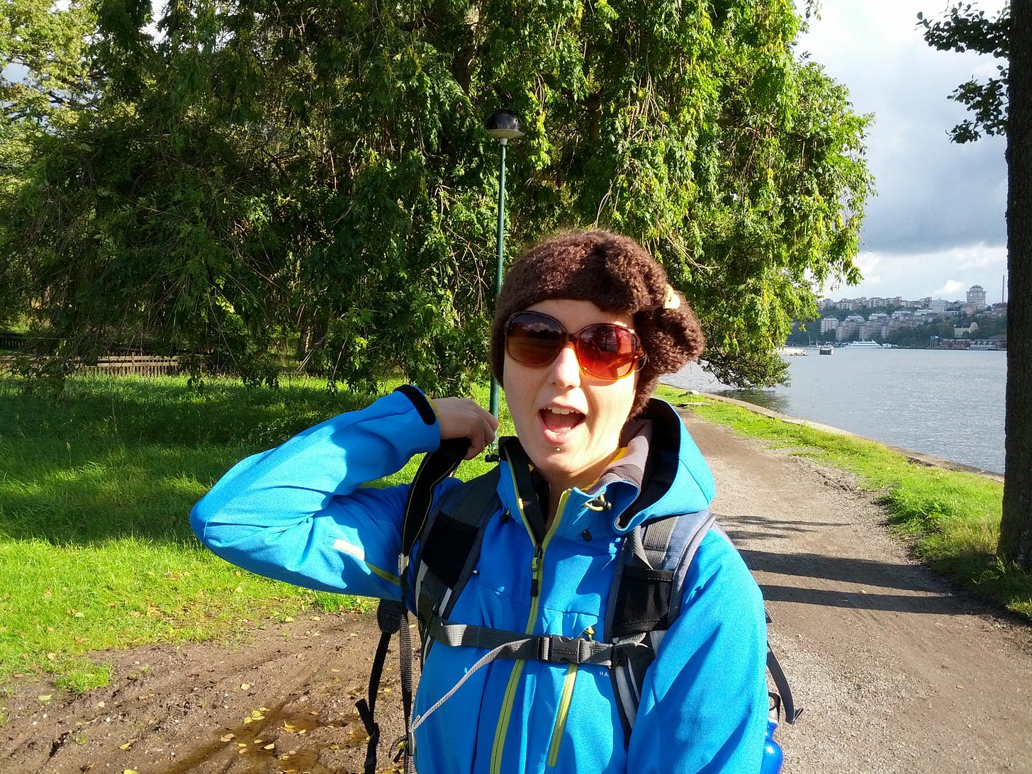 The photo shows the author, a white woman with short brown hair outside. She is wearing a bright blue jacket, a brown headband, sunglasses, and a backpack as she is standing in a park next to water in Stockholm. The photo shows the author, a white woman with short brown hair outside. She is wearing a bright blue jacket, a brown headband, sunglasses, and a backpack as she is standing in a park next to water in Stockholm.