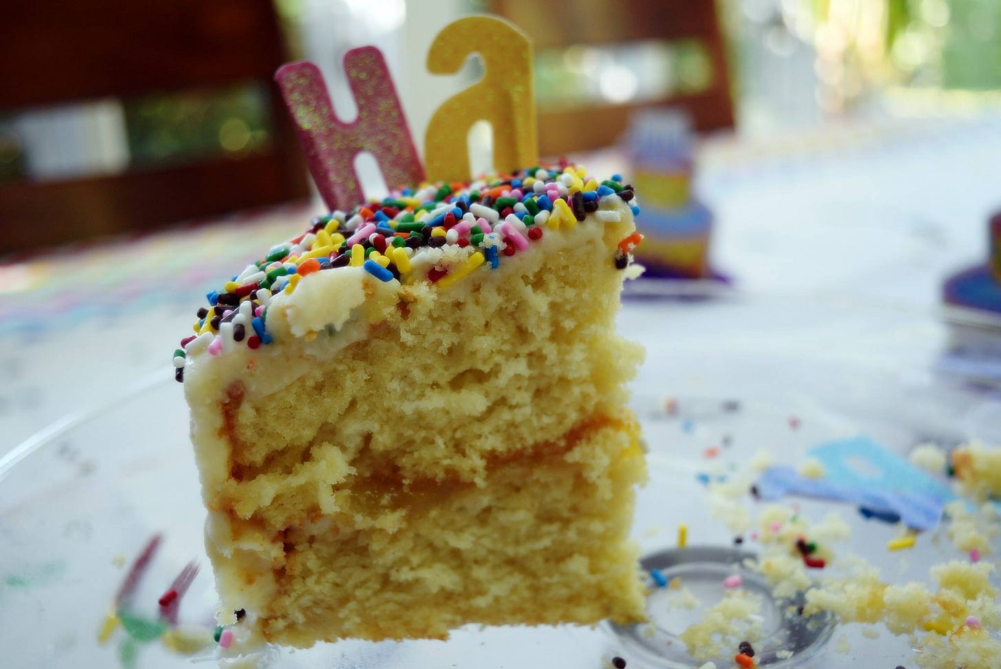 A close-up of a sprinkle-covered slice of birthday cake with the candle letters “Ha” on top, sitting on a crumb-covered plate.
