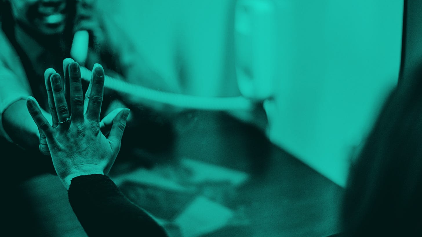 A teal-tinted image of an incarcerated woman and a visitor pressing their hands against the glass.