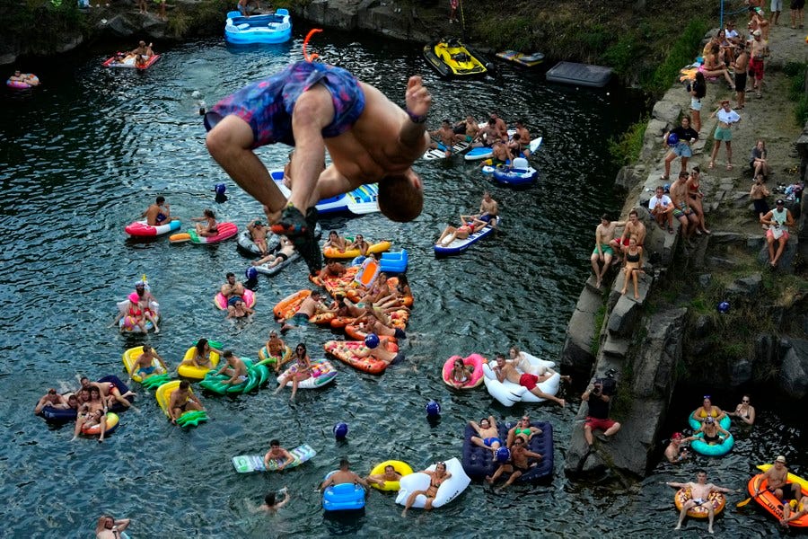 Dozens of people float and play in water, looking up toward a person in mid-air during a high dive. Dozens of people float and play in water, looking up toward a person in mid-air during a high dive.