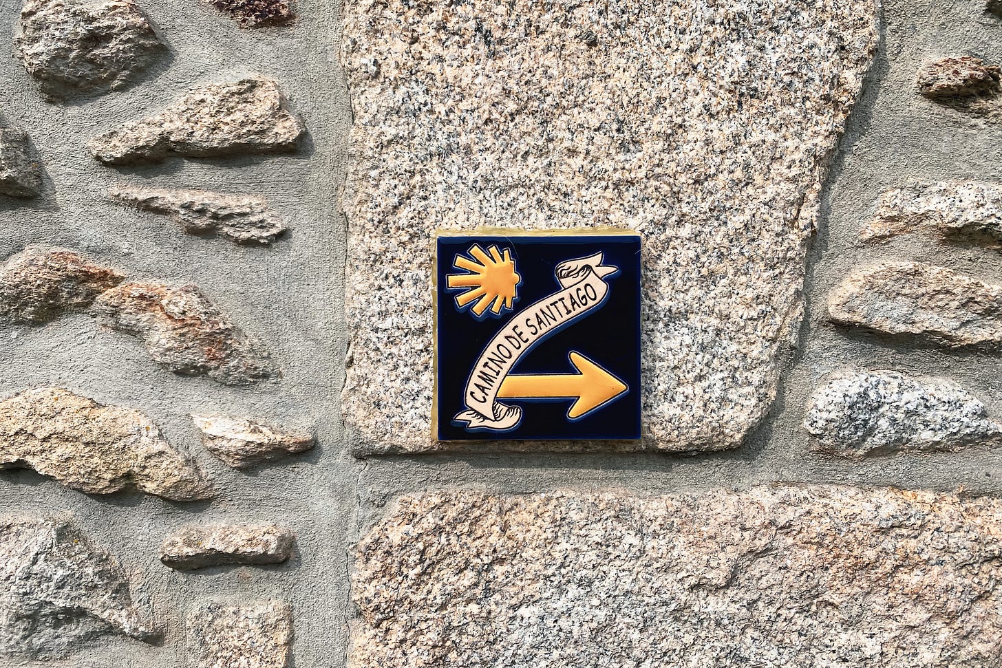 Fotografia de uma parede de pedras com um azulejo onde está escrito "Camino de Santiago" no centro. Ao redor, há uma concha, símbolo do Caminho, e uma flecha indicando a direita. Fotografia de uma parede de pedras com um azulejo onde está escrito "Camino de Santiago" no centro. Ao redor, há uma concha, símbolo do Caminho, e uma flecha indicando a direita.