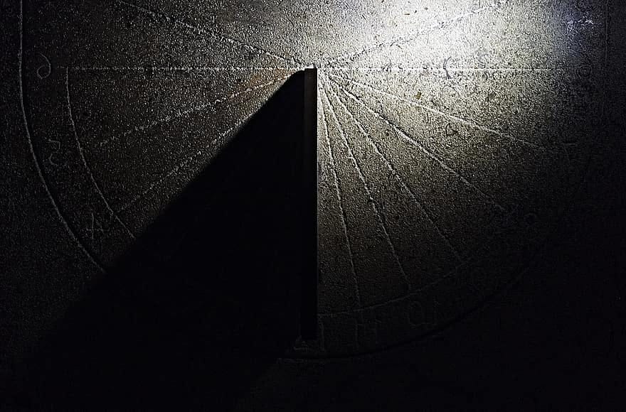 A sundial cast in darkness with a single sharp beam of light creating a shadow across its face