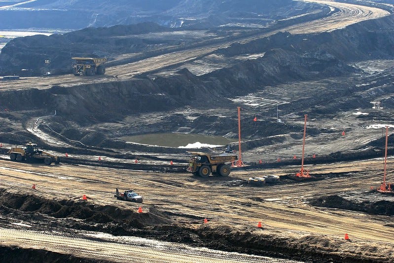 Photo of huge mining dump trucks at work in an open pit mine in Canada's Tar Sands, with heaps of black, churned-up oily earth and ponds of dirty water. Photo of huge mining dump trucks at work in an open pit mine in Canada's Tar Sands, with heaps of black, churned-up oily earth and ponds of dirty water.