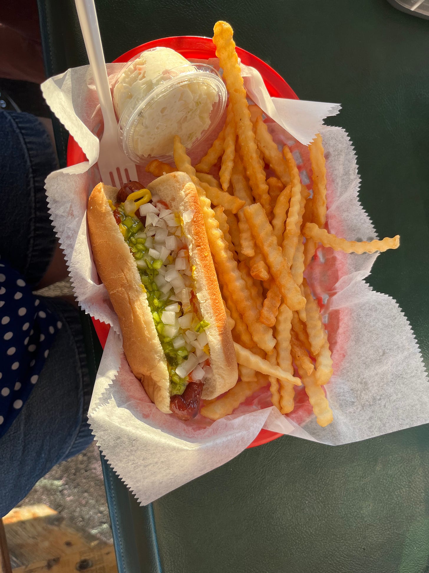 Picture shown from above of a red basket full of fries, a grilled hot dog with onions and pickle relish and mustard, and cole slaw! A fork is stuck in the side of the basket as if someone's about to take a delicious bite.