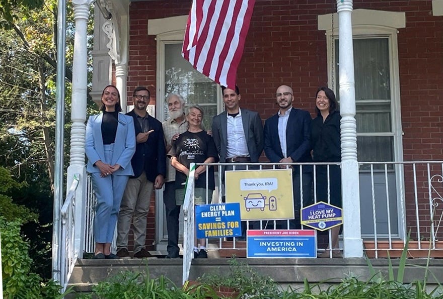Seven people pose for a photo on the porch of a brick house with white wooden pillars and scrollwork. Signs on the porch railing read 'Clean energy plan = savings for families,' 'I love my heat pump,' 'President Joe Biden: Investing in America," and 'Thank You IRA' — the last with a cartoon of a smiling car plugged into an EV charger