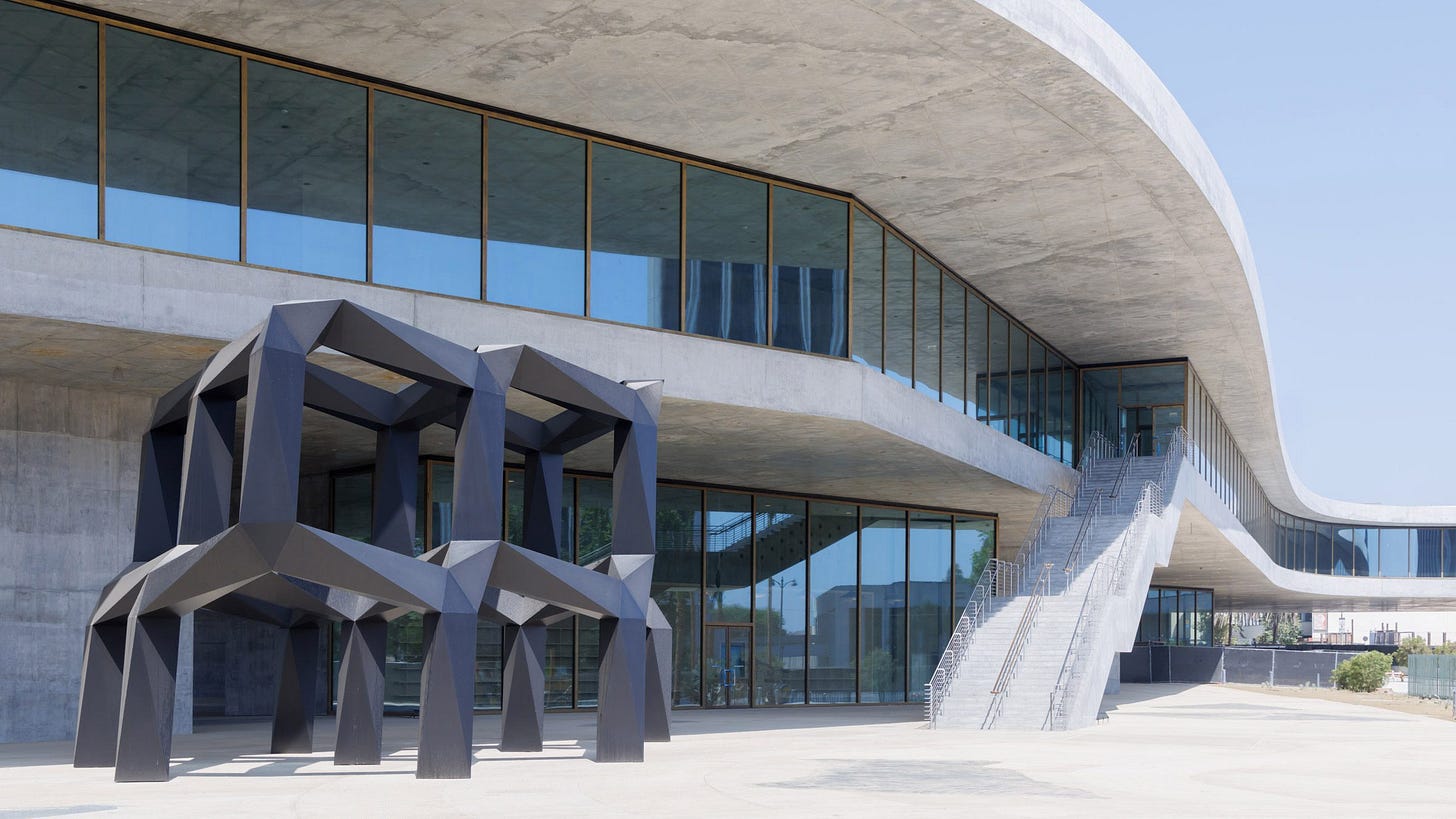 The exterior of the Geffen Galleries with a sweeping curved concrete roofline, glass facade, and a large angular sculpture installed near the entrance. The exterior of the Geffen Galleries with a sweeping curved concrete roofline, glass facade, and a large angular sculpture installed near the entrance.
