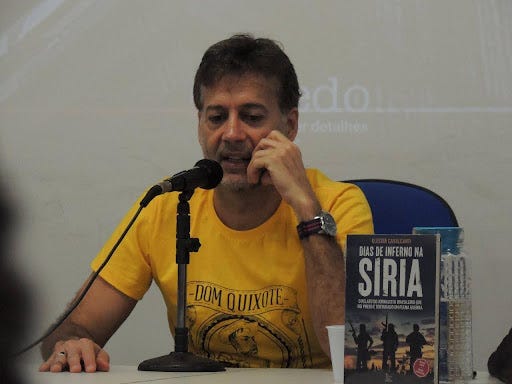 Foto de homem branco de camiseta amarela falando ao microfone ao lado de um livro sobre a Síria