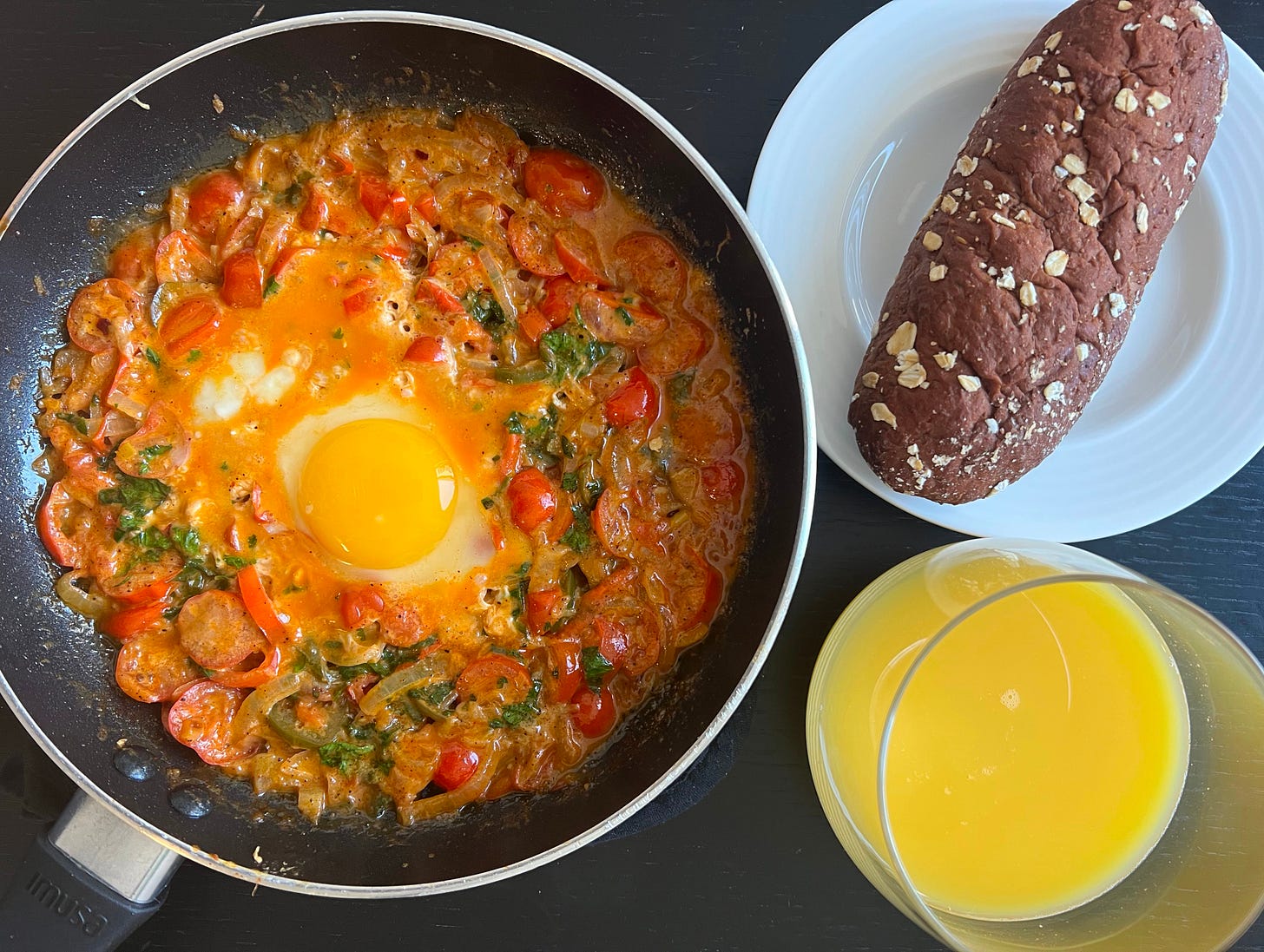 A homemade shakshuka by Liat Portal for Foodie Disorder, featuring a rich tomato sauce with bell peppers and a golden egg yolk nestled in the center. A white plate holds a rustic brown bread roll, and a glass of orange juice sits on the side. This meal, photographed for The Liat Show, highlights the comforting simplicity of shakshuka as a staple dish.