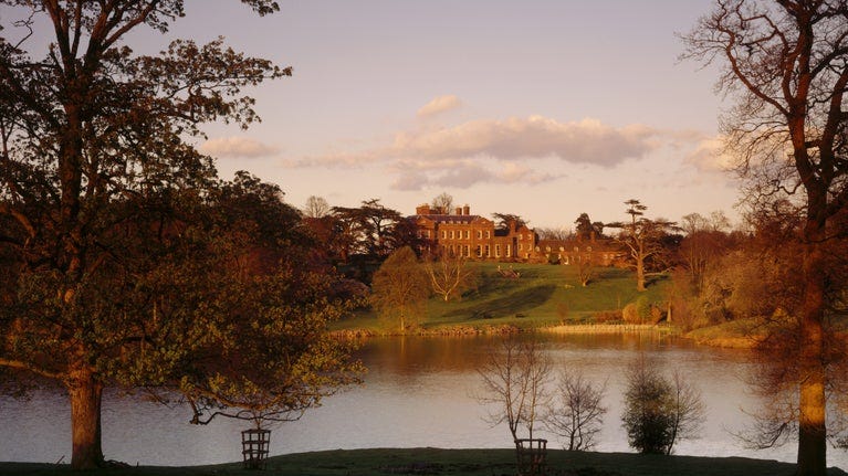 The West Front of Dudmaston in Shropshire from across the Big Pool at sunset. The 8-acre garden is sloping to the lake from the red brick houses which include the main block, domestic range and Brew House.