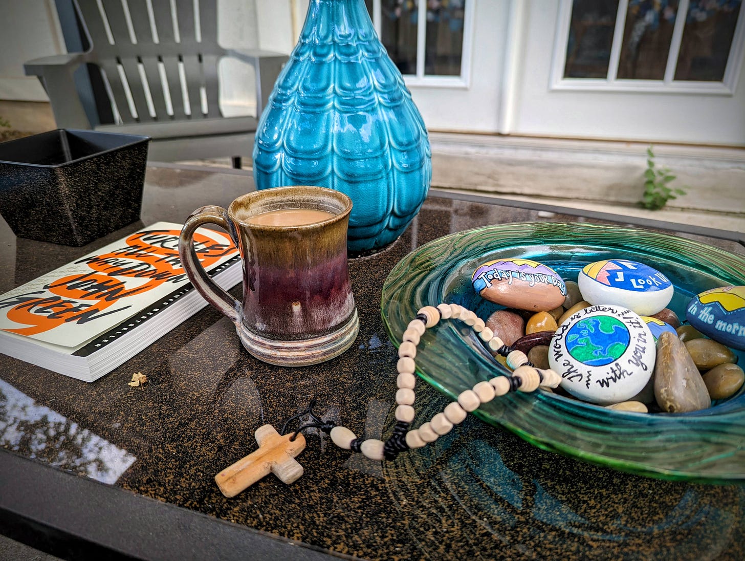 A picture of items on a table outside. Items include a book, cup of coffee, a bowl with decorative rocks, and a blue vase. In the foreground are a set of prayer beads with a cross.