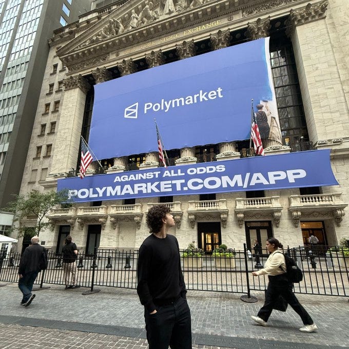 Facade of the New York Stock Exchange building with classical columns and steps. Large blue banners display Polymarket logo and text All Odds and polymarket.com/app. American flags hang on either side. People including a man in black shirt looking up and a woman in beige walking nearby. Plants and barriers visible in foreground.