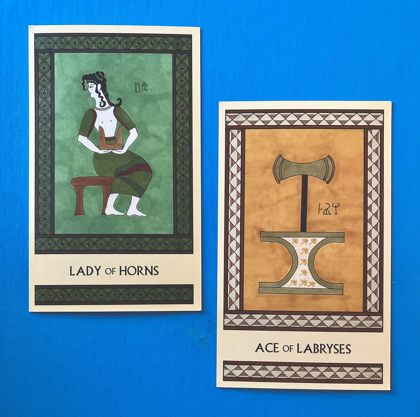 Two Minoan Tarot cards side by side on a bright sky blue background. The Lady of Horns is painted in shades of green and brown. It shows a Minoan woman seated on a small stool, facing right but looking left, holding a pair of sacred horns in front of her. The Ace of Labryses is painted in shades of golden yellow, cream, and tan. It shows a large gold labrys standing atop a small altar with incurved sides. 