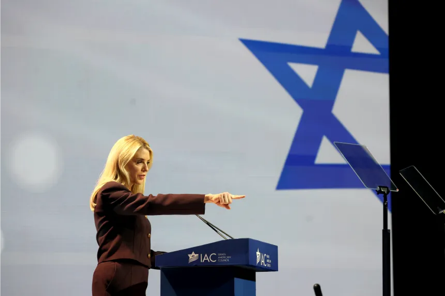 Pam Bondi, standing before the flag of the state whose intelligence lineage she now protects more fiercely than Epstein’s victims.