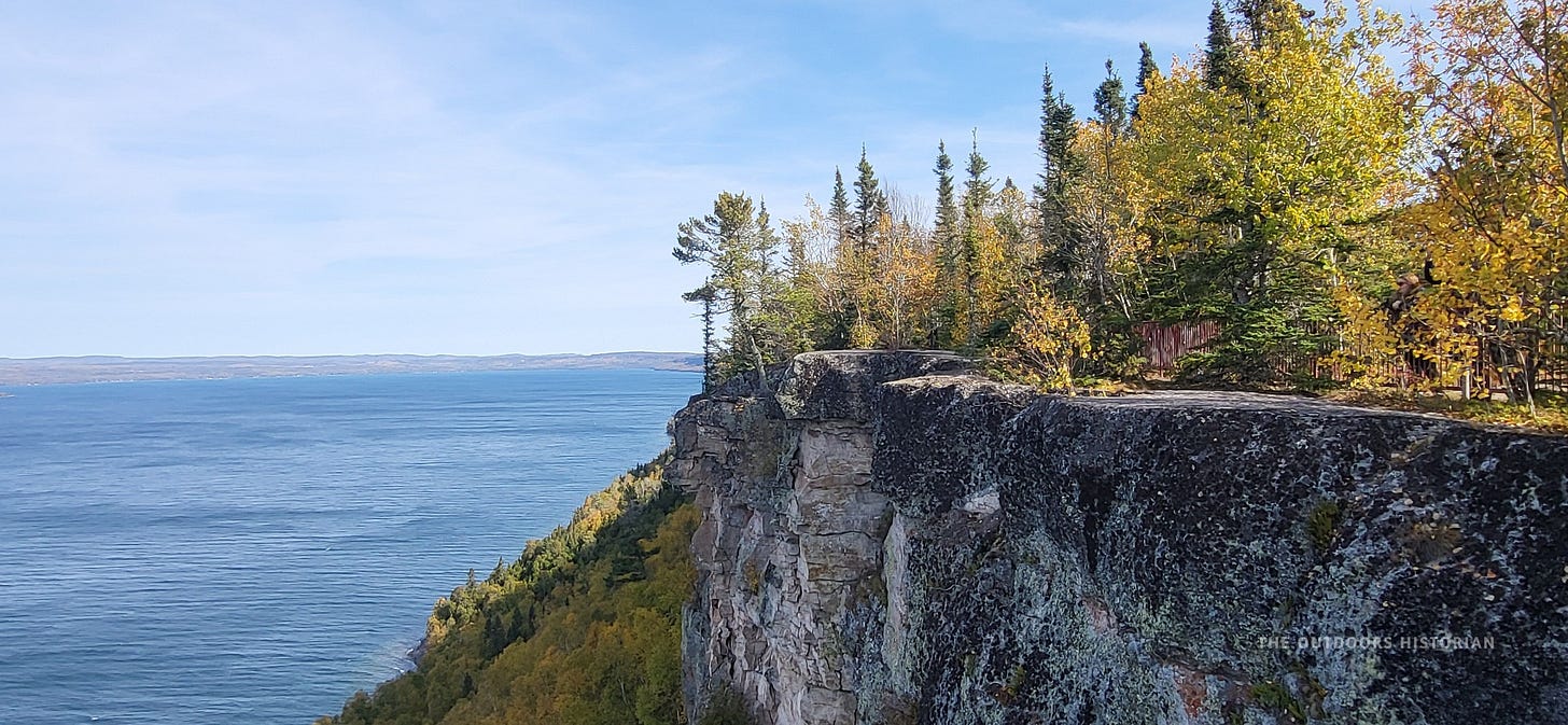 A view looking straight down a sheer, vertical rock cliff face. The rock is dark and jagged, dropping down to a dense green forest and the blue lake below.