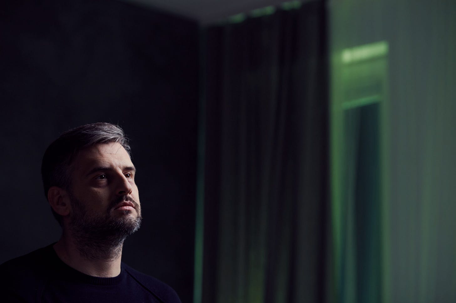 A man with greying hair stares pensively up in a dark room.