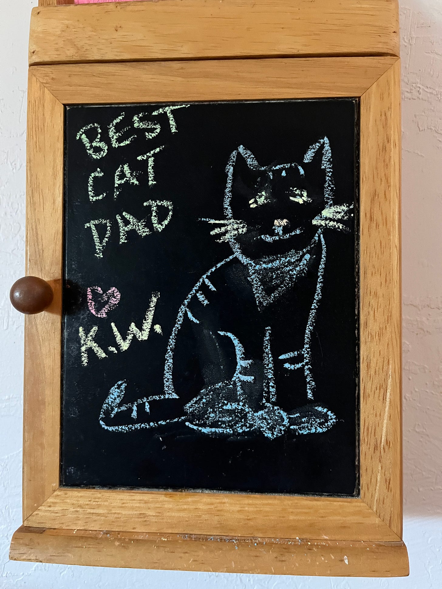 photo of a wooden cabinet with a chalkboard front. The chalkboard shows a rough image of a smiling tabby cat with the caption "BEST CAT DAD [Heart] K.W."